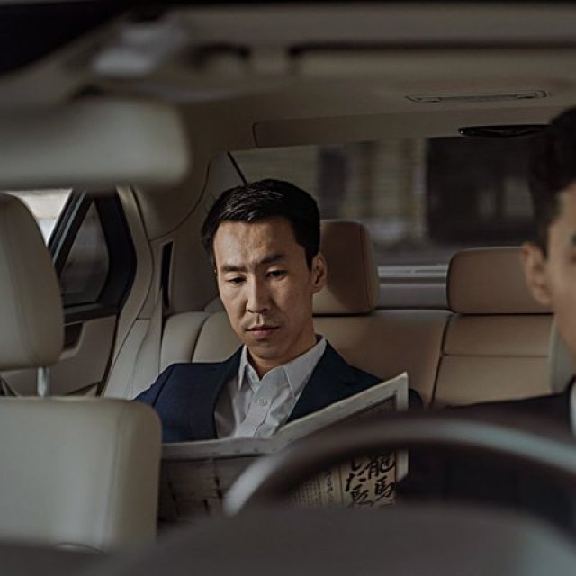 A passenger reads in the rear seat of a chauffeur-driven saloon, the chauffeur at the wheel ahead.