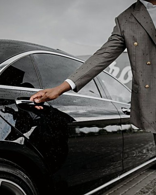 A chauffeur in a tailored suit opens the rear door of a black saloon beaded with rain.