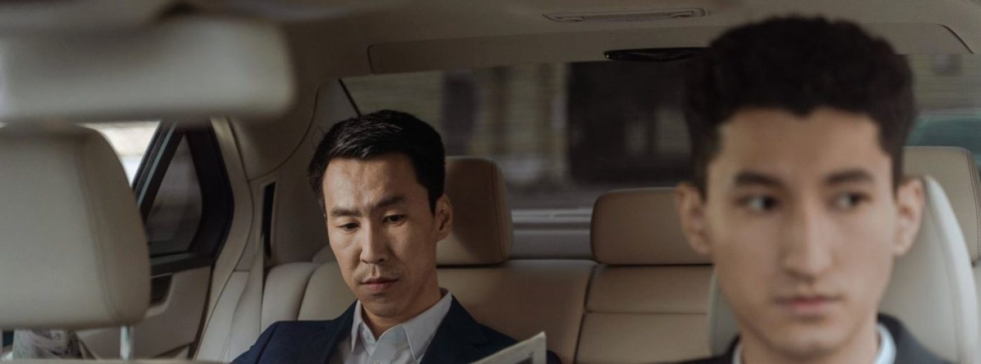 A passenger reads a newspaper in the rear seat while the chauffeur drives.