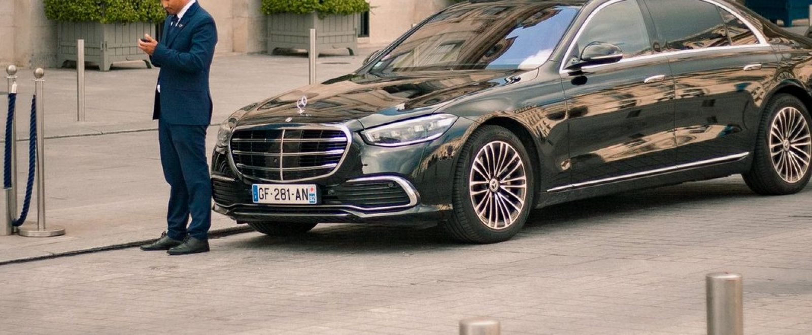 A chauffeur in a navy suit stands beside a black Mercedes-Benz S-Class on a hotel forecourt.